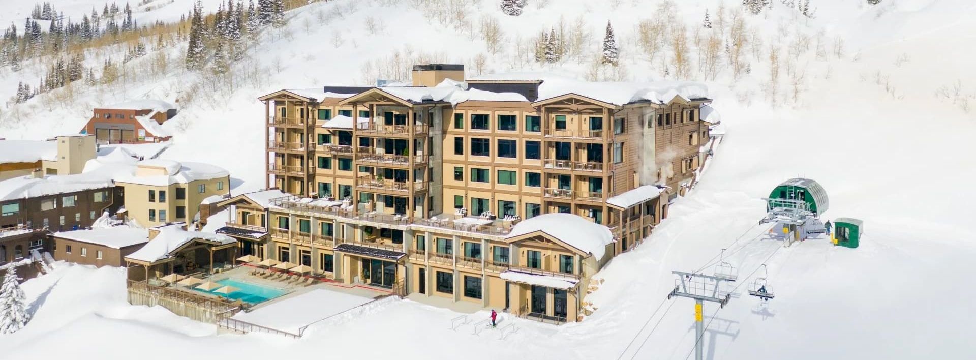 A snowy alpine ski resort with a multi-story hotel, chairlift cables, and mountain peaks under a blue sky.