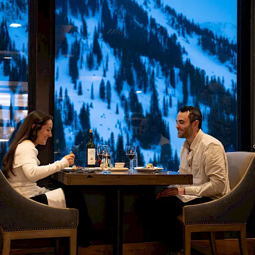 Two people dine at a table by a window, clinking drinks while watching snow-covered mountains outside.