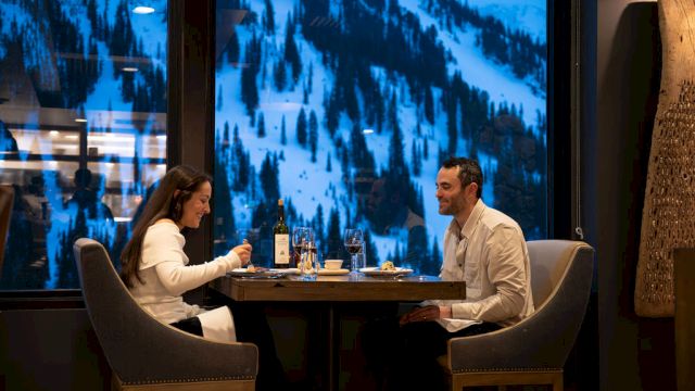 Two people dine at a table by a window, clinking drinks while watching snow-covered mountains outside.