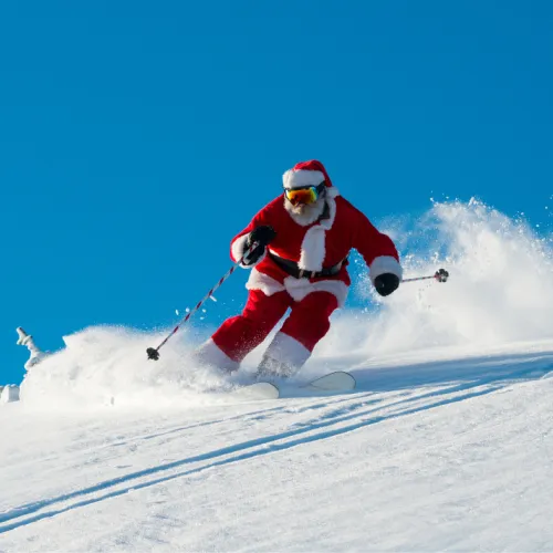 Santa Claus in a red suit skis down a snowy mountain, sending up a big splash of powder against a clear blue sky.