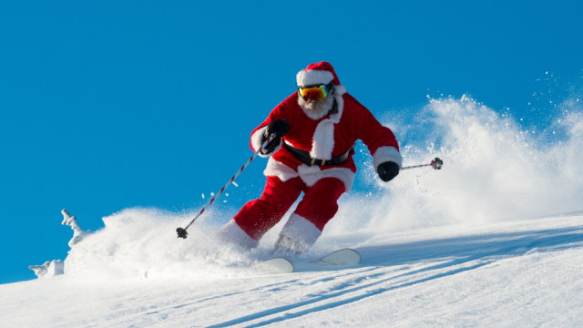 Santa Claus in a red suit skis down a snowy mountain, sending up a big splash of powder against a clear blue sky.