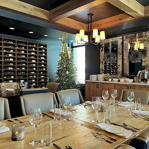 A dressed restaurant scene with a decorated Christmas tree, glassware at tables, and wall-to-wall wine bottles in shelves.