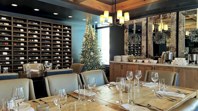 A dressed restaurant scene with a decorated Christmas tree, glassware at tables, and wall-to-wall wine bottles in shelves.