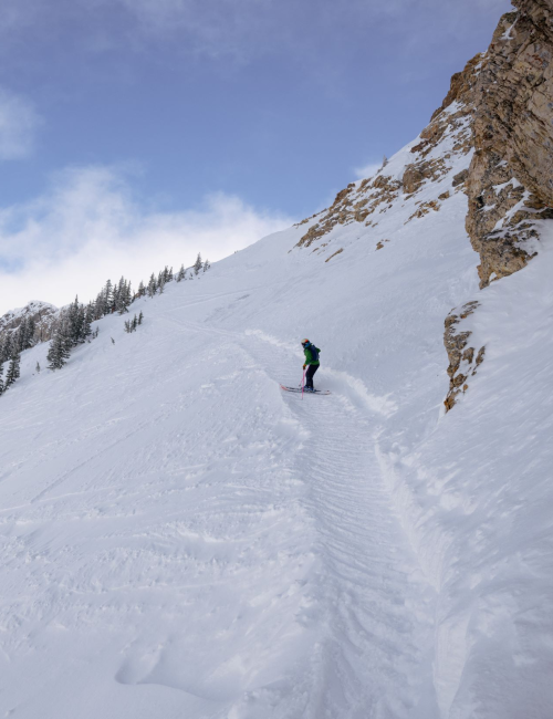 A skier carves down a snowy mountain slope beside rock cliffs, with pine trees and clouds in the bright sky behind.